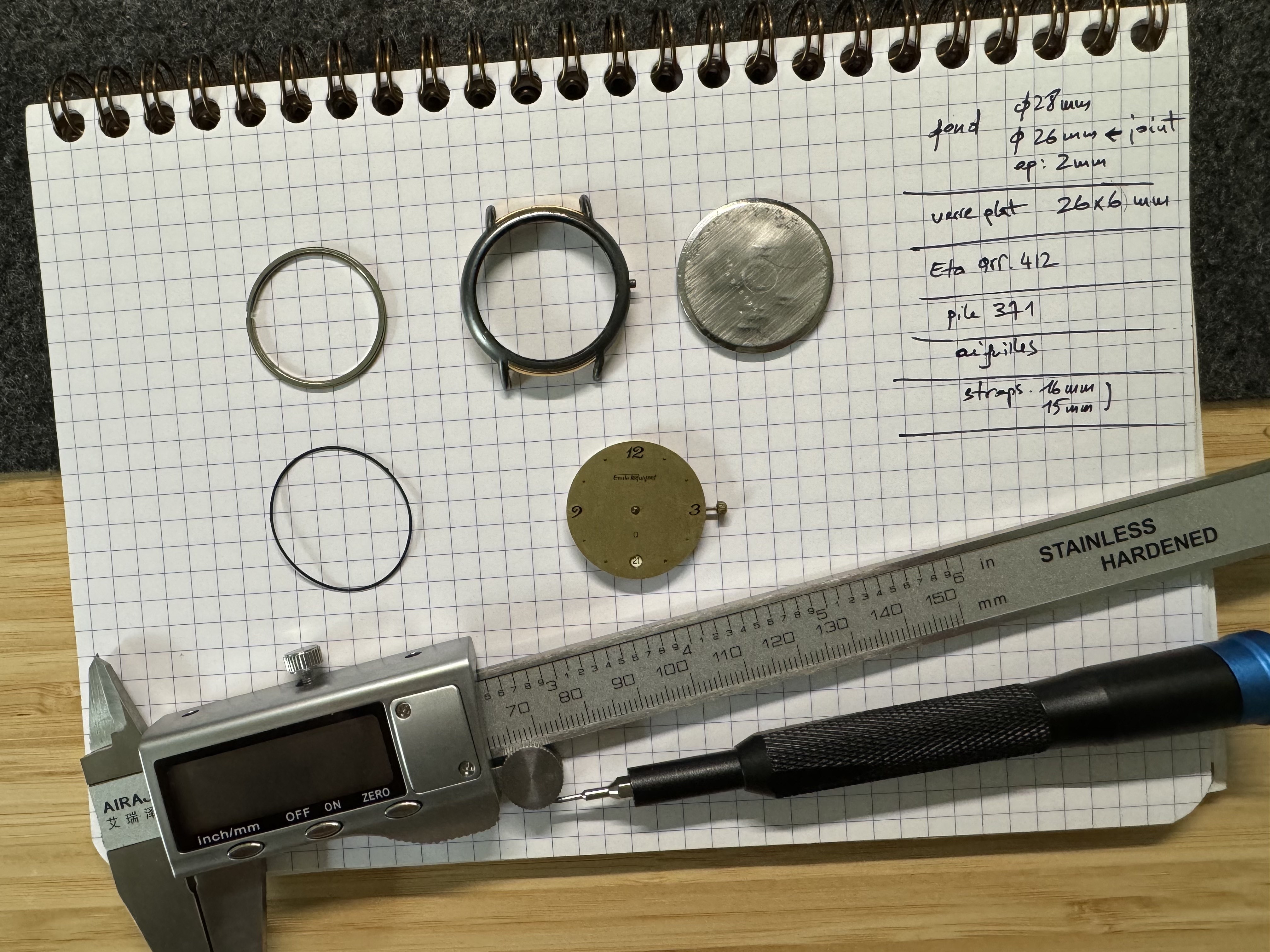 Disassembled Emile Pequignet watch components — case, caseback, crystal, dial and gasket — alongside a digital caliper and handwritten repair notes