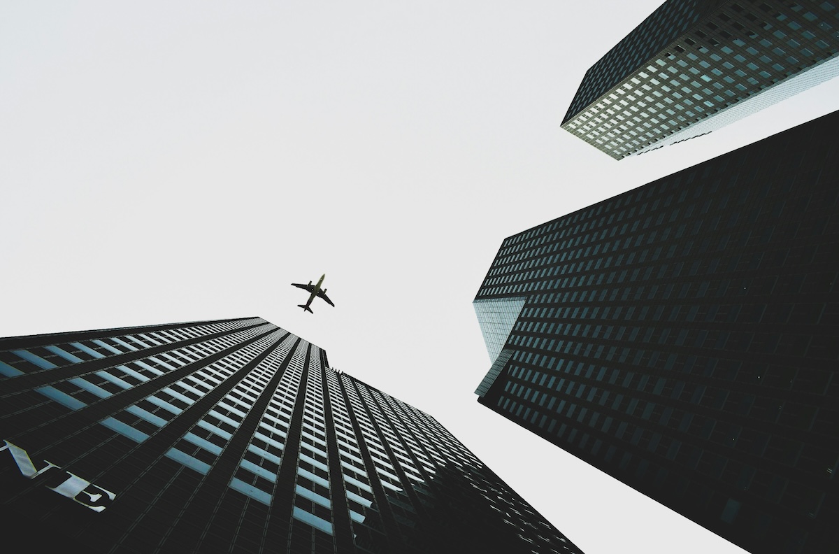Looking straight up between three dark glass office towers into a pale sky, with a small plane passing high above the gap between them